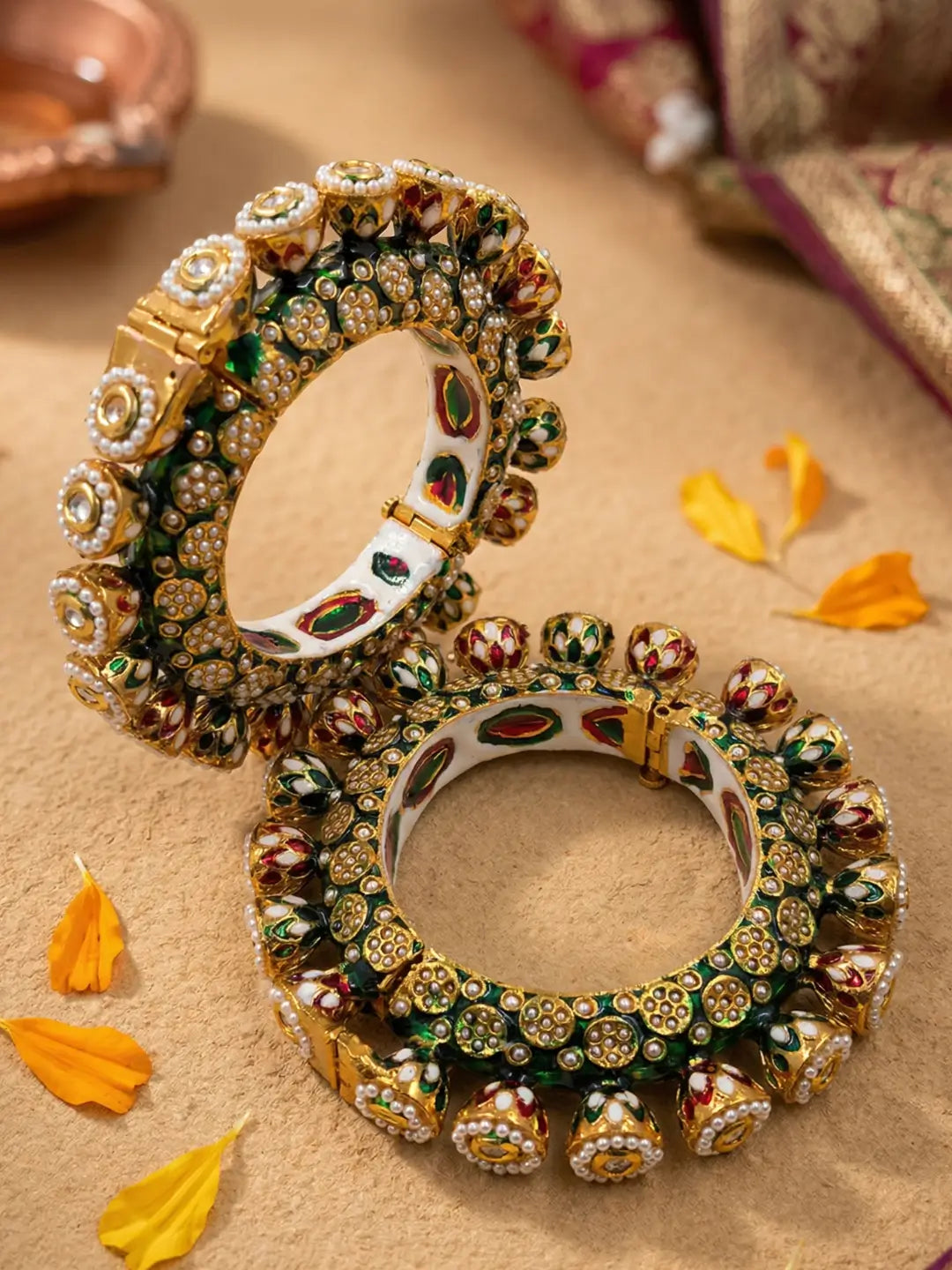 Two gold-plated Indian Kundan bangles displayed on a textured beige surface with scattered yellow flower petals. The bangles feature intricate green enamel work with white pearls and kundan stones 