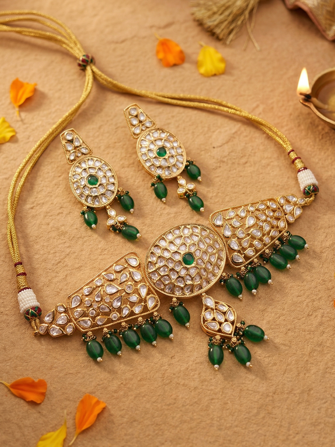 Gold-plated traditional Indian choker jewelry set featuring a necklace, earrings, and a central pendant, embellished with clear polki stones and green beads, arranged on a textured beige surface with scattered orange and yellow petals.