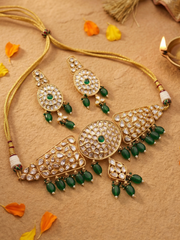 Gold-plated traditional Indian choker jewelry set featuring a necklace, earrings, and a central pendant, embellished with clear polki stones and green beads, arranged on a textured beige surface with scattered orange and yellow petals.