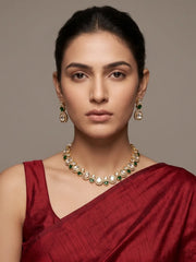 A woman wearing a gold-plated Kundan necklace and matching drop earrings with green stones paired with a deep red silk saree, set against a solid taupe background.
