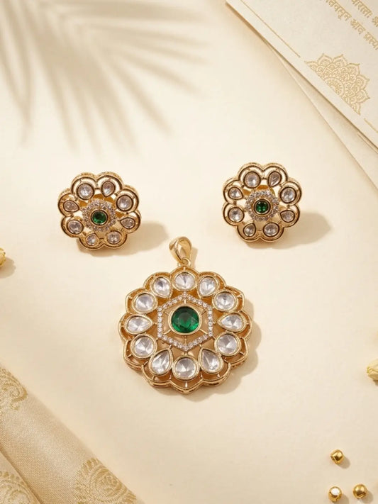 Gold-toned traditional Indian Kundan jewelry set featuring a pendant and matching stud earrings with clear stones and a central green stone, displayed on a cream-colored surface.