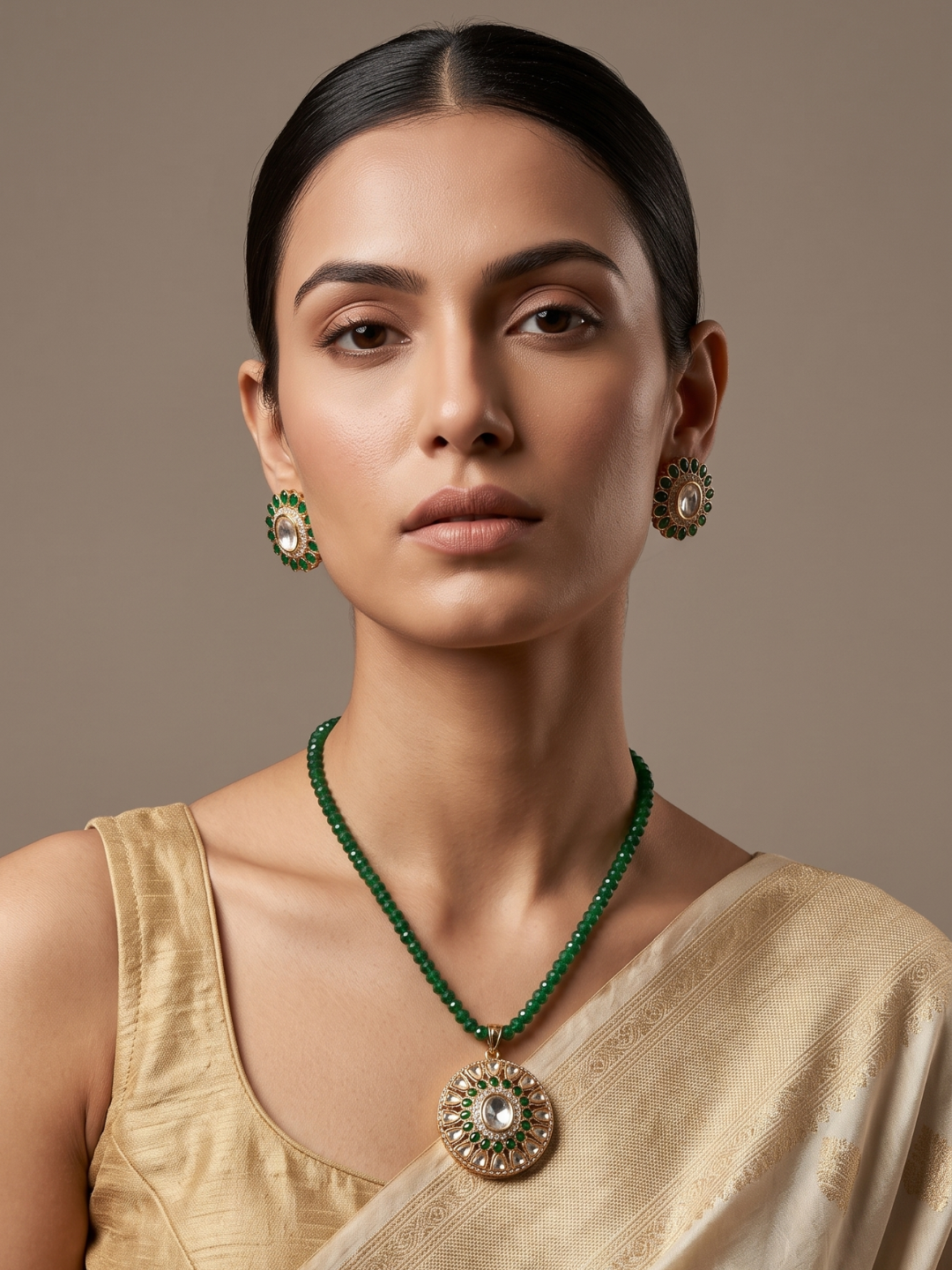 A woman wearing a green beaded necklace with a large ornate gold pendant and matching green and gold earrings, styled with a beige saree against a neutral brown background.
