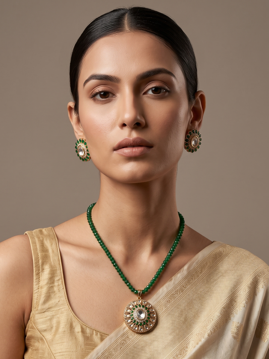 A woman wearing a green beaded necklace with a large ornate gold pendant and matching green and gold earrings, styled with a beige saree against a neutral brown background.