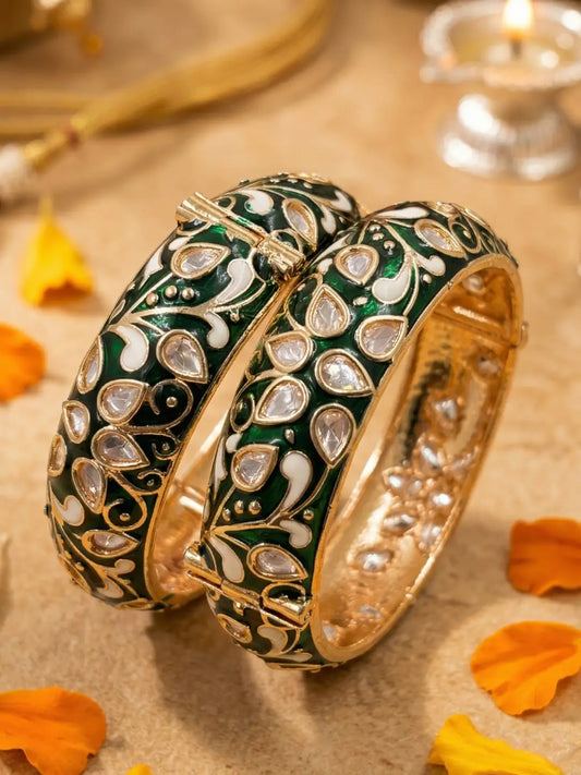 Two ornate gold-plated bangles decorated with dark green enamel, white floral motifs, and inlaid clear stones, displayed on a beige surface with orange flower petals and a softly blurred background.