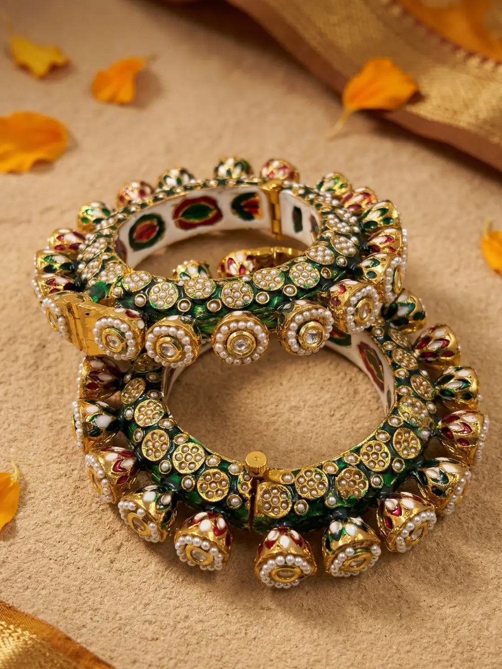 A stack of three ornate Indian Kundan bangles with intricate gold-plated detailing, green enamel, and red floral accents, displayed on a textured beige surface with scattered orange flower petals.