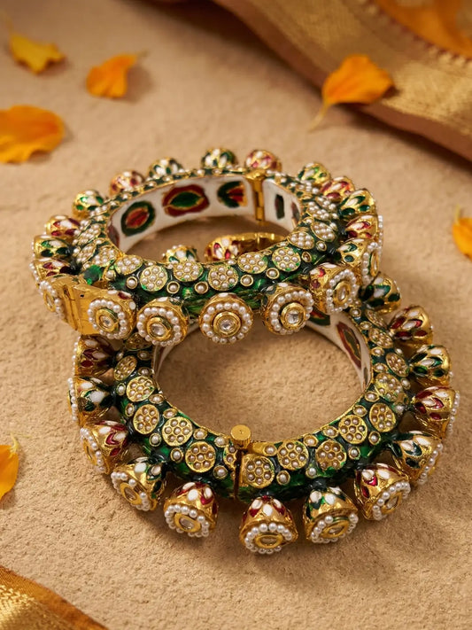 A stack of three ornate Indian Kundan bangles with intricate gold-plated detailing, green enamel, and red floral accents, displayed on a textured beige surface with scattered orange flower petals.