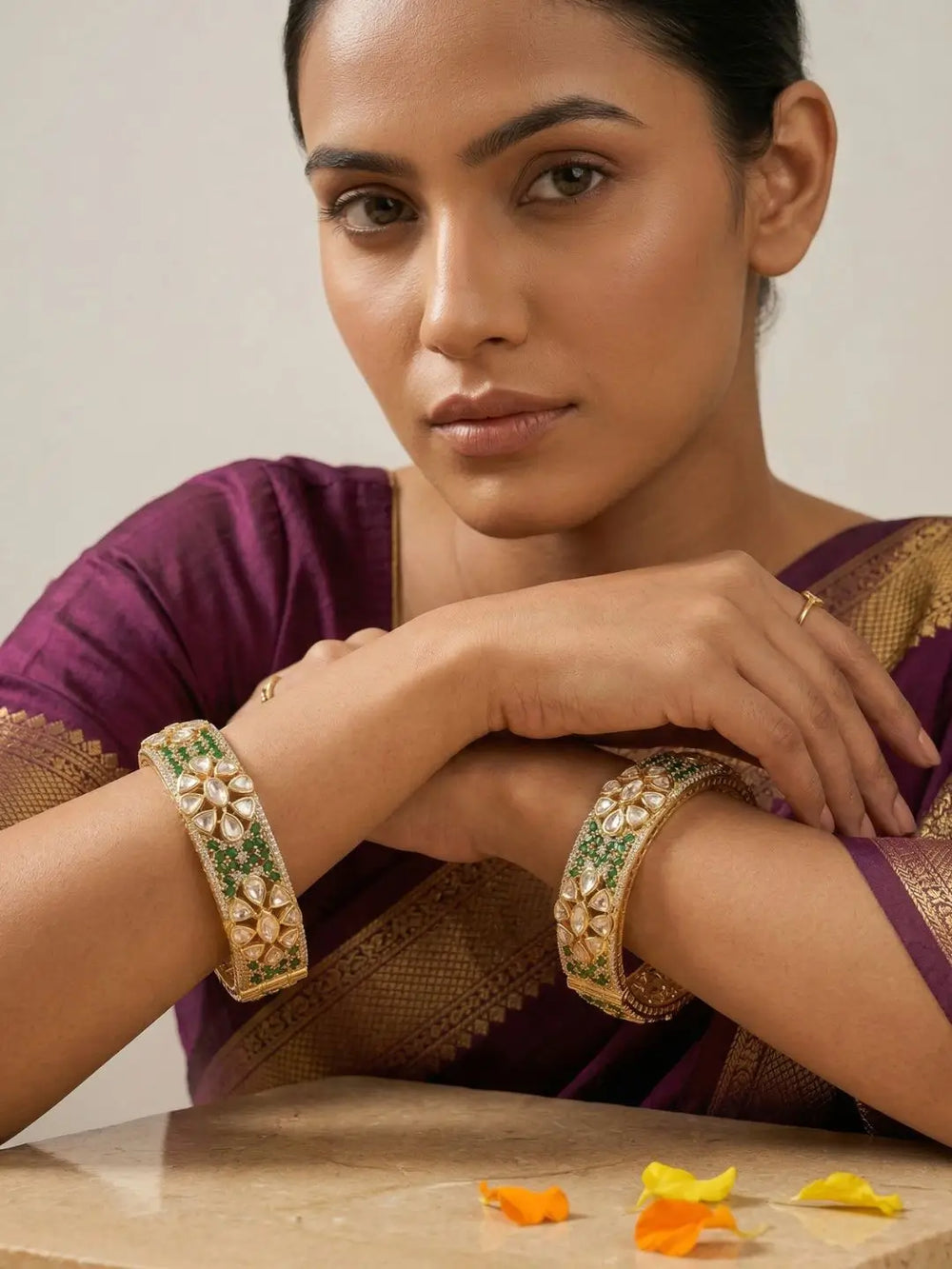 A woman wearing ornate gold-plated green emerald, kundan and polki-studded bangles on her both wrists posed in a purple saree against a neutral background.