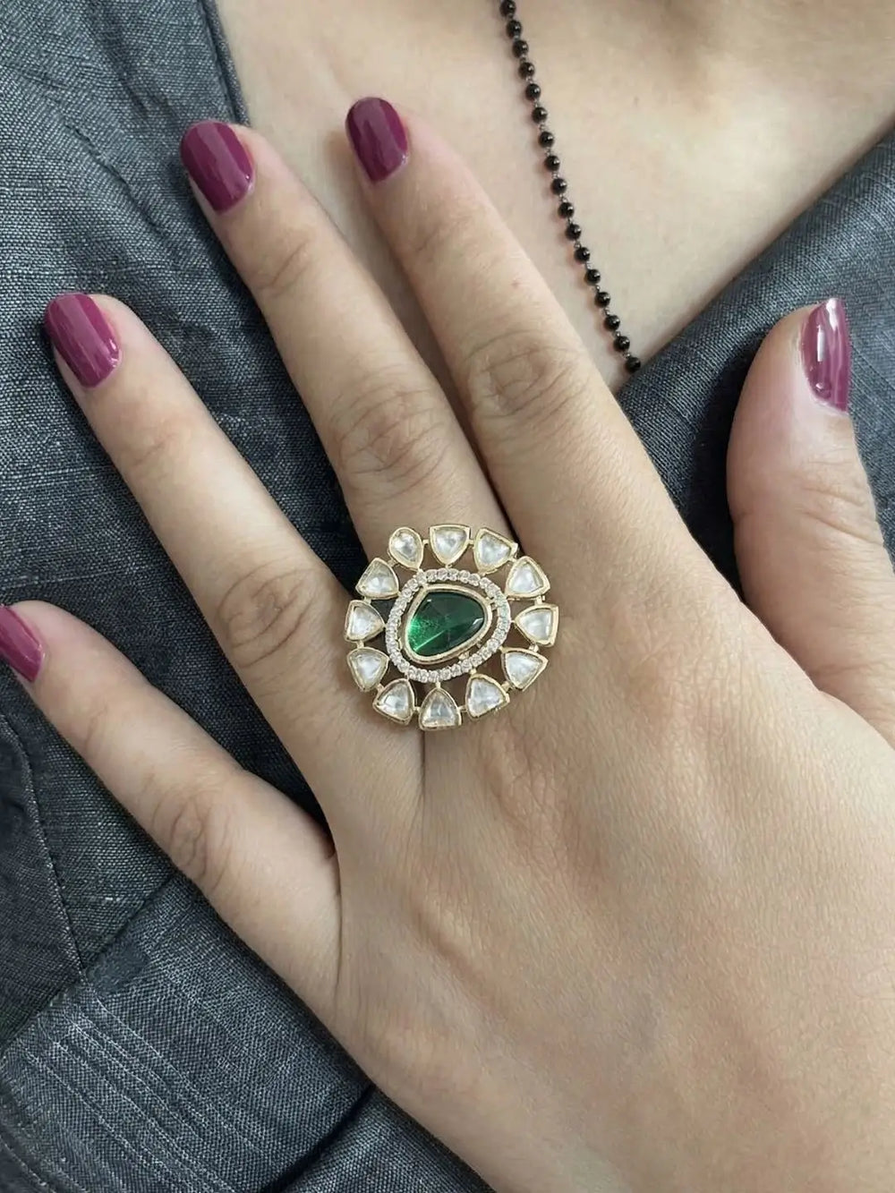 A hand wearing a Kundan ring with a green gemstone on a gray fabric background