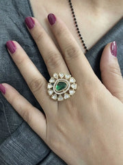 A hand wearing a Kundan ring with a green gemstone on a gray fabric background