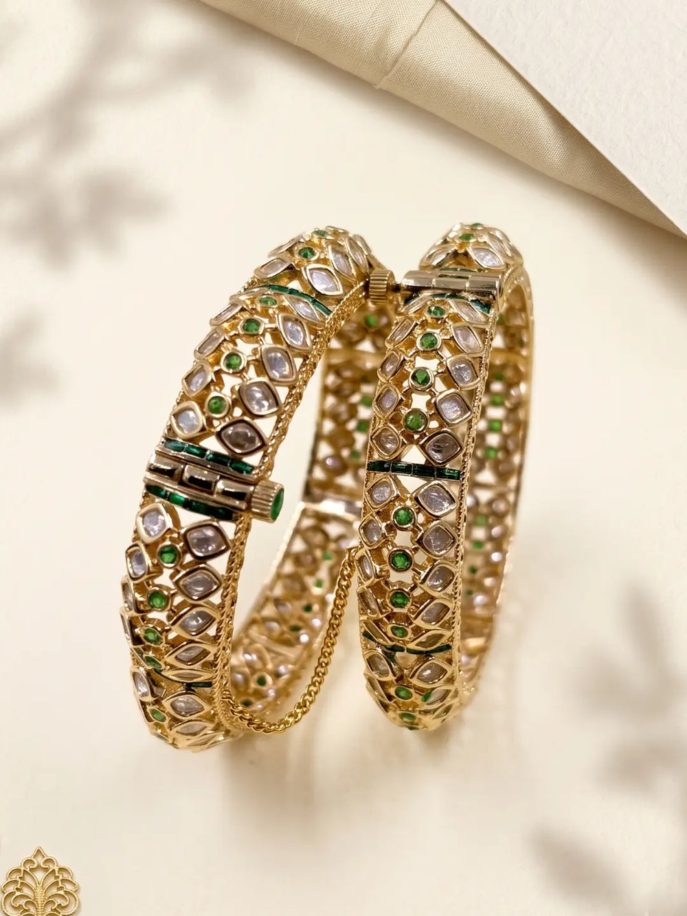 A pair of ornate, gold-toned green uncut polki kada bangles decorated with clear polki stones, displayed on a light cream background.