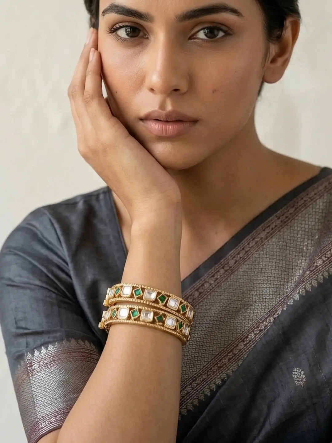 A close-up of a woman wearing a dark gray saree and stacked gold-toned bangles with green and white stones, posing with her hand on her cheek against a neutral light background.
