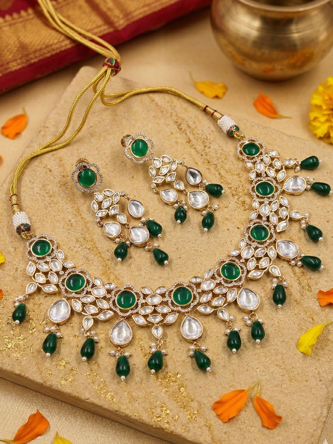 Ornate gold plated kundan necklace and earringg set with emerald green and white polki stones on a textured surface with flowers.