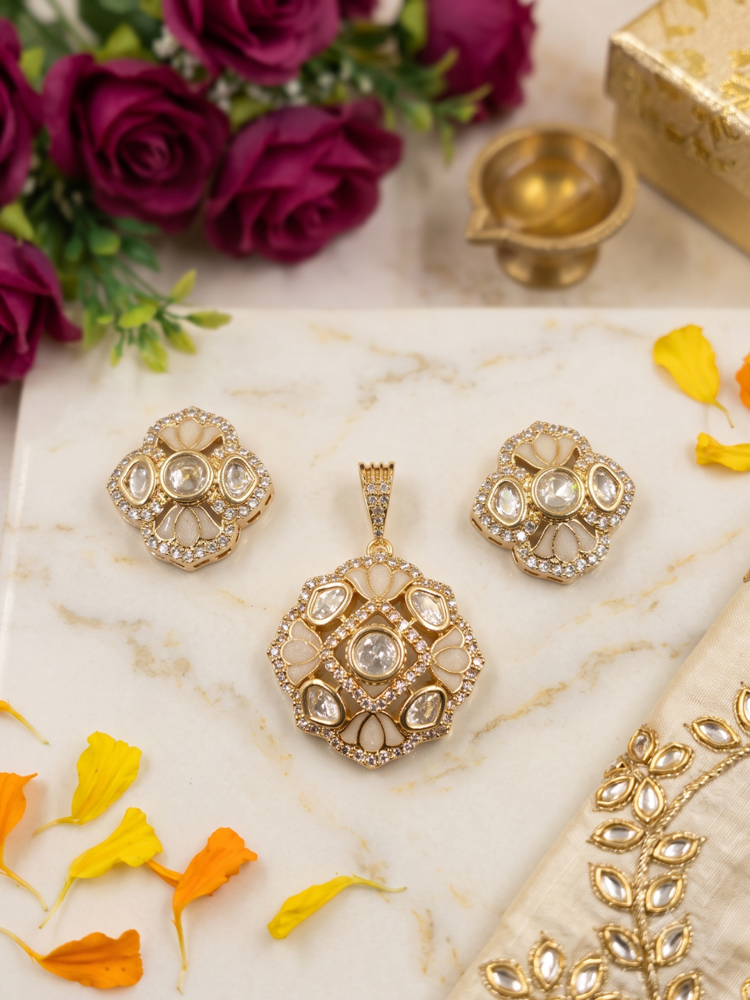 Gold-plated traditional Indian pendant and earring set featuring a matching pendant, earrings, and a decorative fabric piece, arranged on a white marble surface with rose petals, flowers, and a diya in the background.