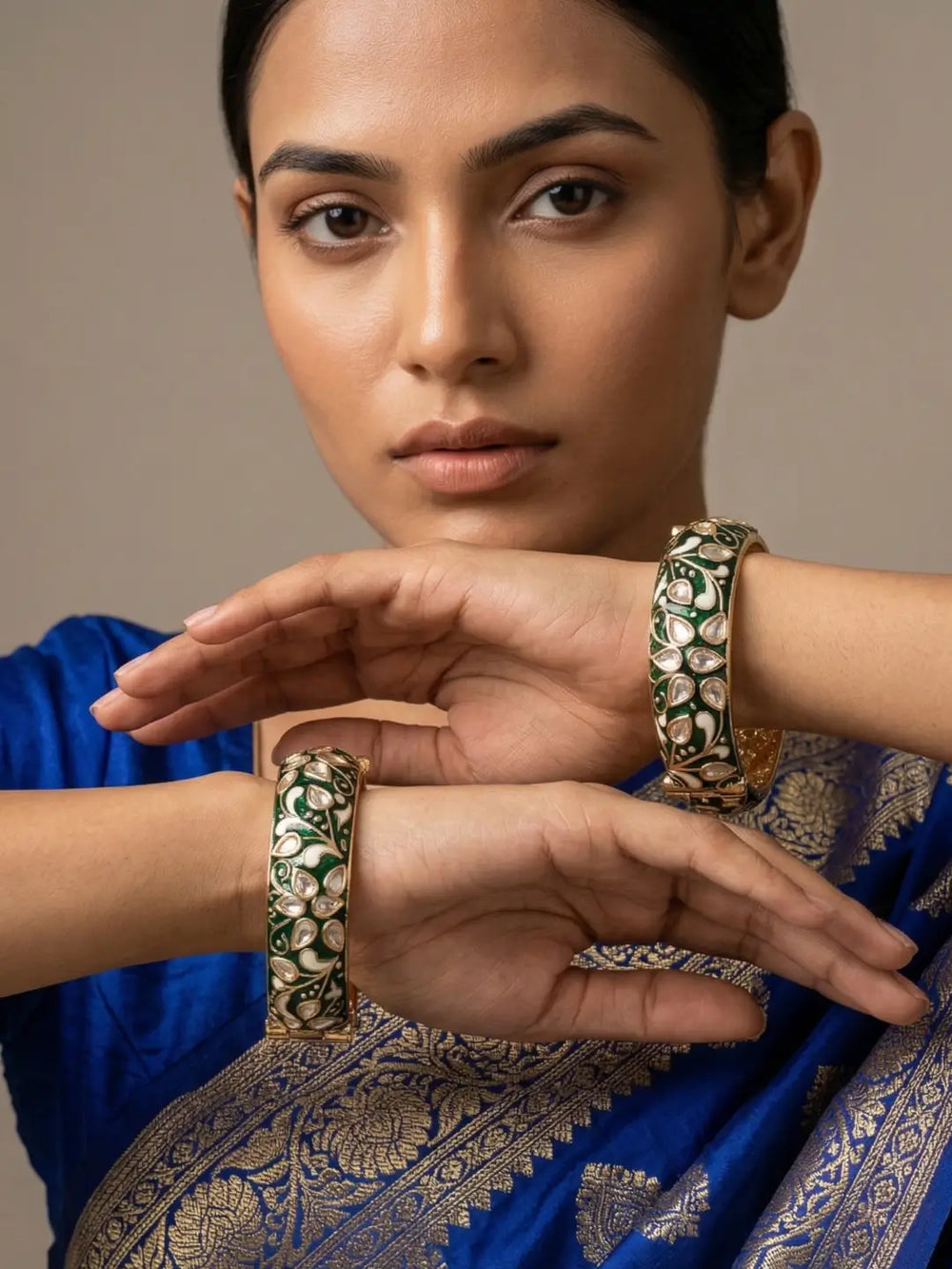 A close-up of a woman wearing ornate gold bangles with green and white floral meenakari paired with a royal blue sari featuring intricate gold zari work.