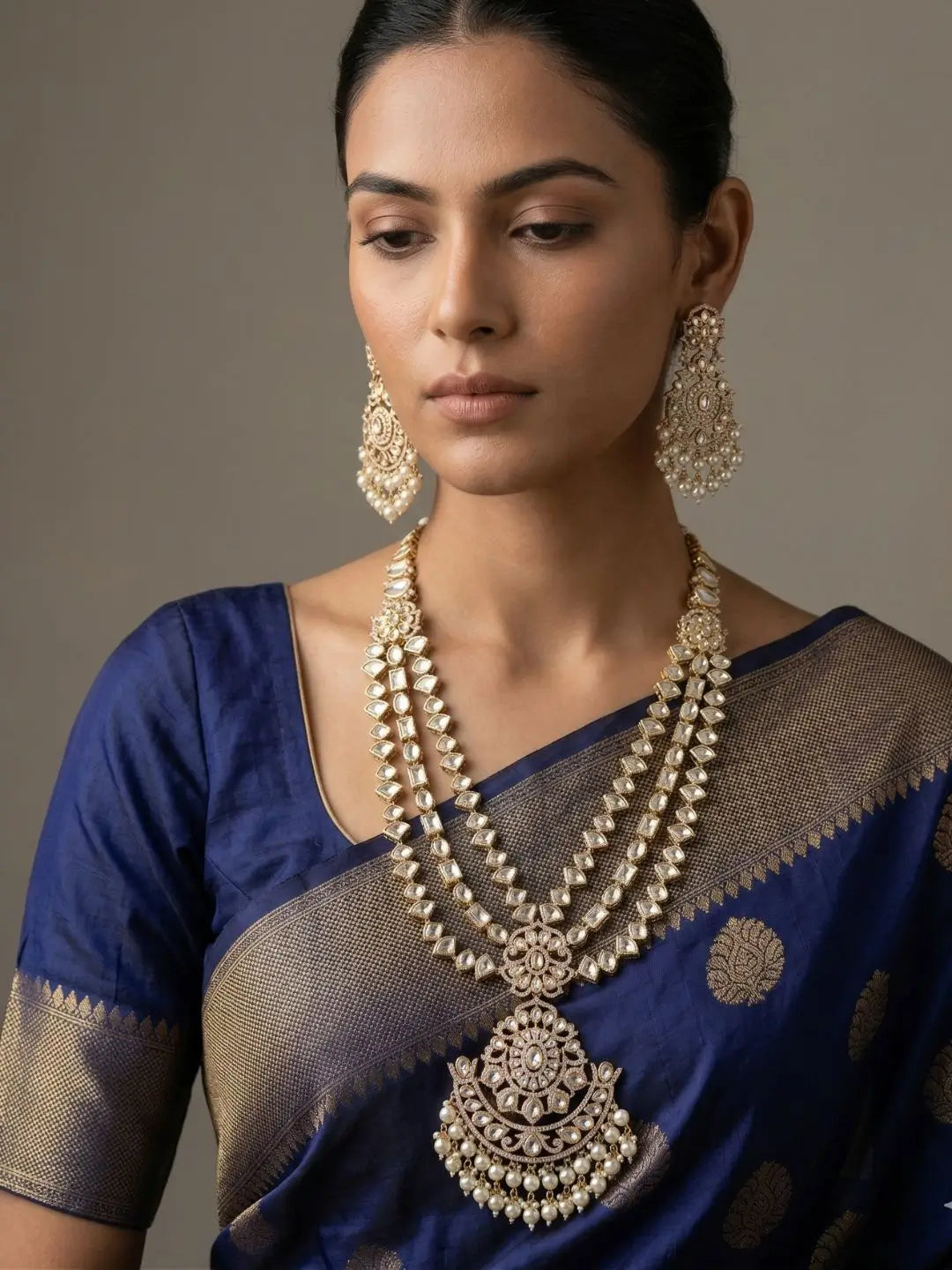 A woman wearing an ornate traditional gold-plated long layered Kundan polki necklace and matching earrings, paired with a royal blue saree with gold zari work, set against a neutral gray background.