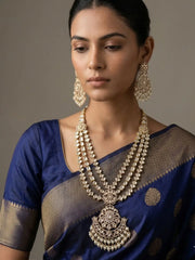 A woman wearing an ornate traditional gold-plated long layered Kundan polki necklace and matching earrings, paired with a royal blue saree with gold zari work, set against a neutral gray background.