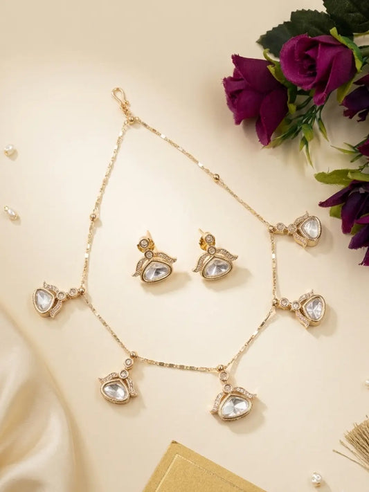 Gold-toned Indian jewelry set featuring a necklace, matching earrings, and a chain, decorated with clear teardrop-shaped stones and small gold beads, styled on a cream background with purple roses.