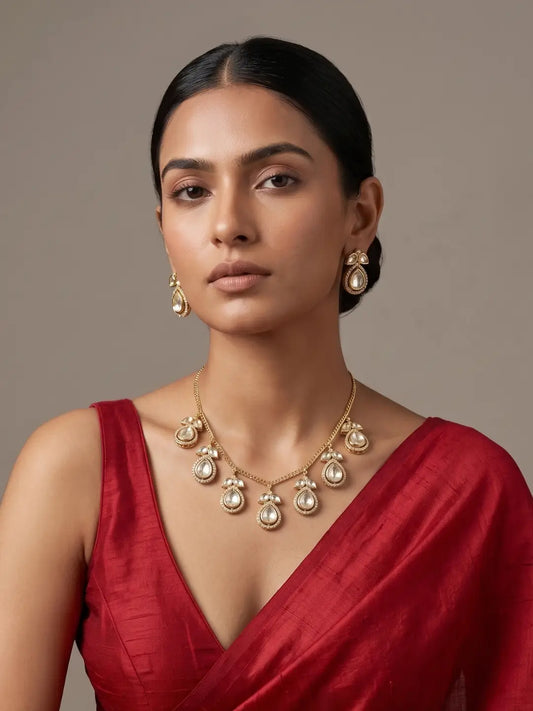 A woman wearing a gold-toned statement necklace and matching drop earrings with polki-like teardrop charms, styled with a deep red sari against a neutral taupe background.