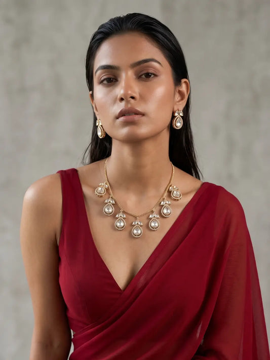 A woman wearing a gold-plated chain statement necklace with polki drops and matching teardrop earrings styled with a deep red draped dress against a textured gray background.