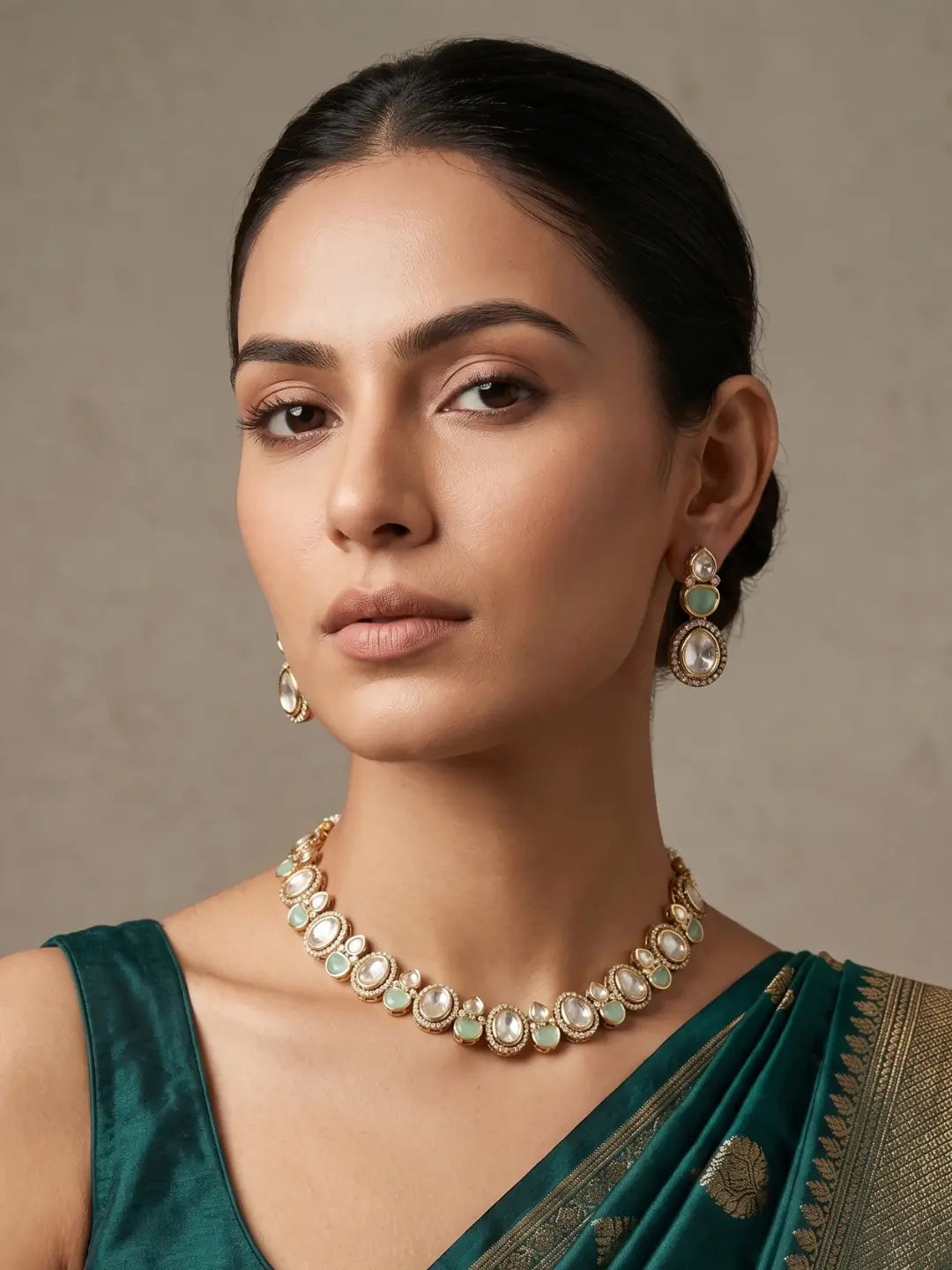 A woman wearing a gold-toned necklace and matching drop earrings with white stones and light green inlays, paired with a dark teal sari against a neutral beige background.