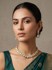 A woman wearing a gold-toned necklace and matching drop earrings with white stones and light green inlays, paired with a dark teal sari against a neutral beige background.