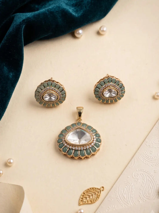 A gold-toned jewelry set featuring a pendant, matching earrings, and a small gold leaf charm, decorated with green enamel and clear stones, arranged on a cream surface with a dark teal velvet cloth and pearl props.