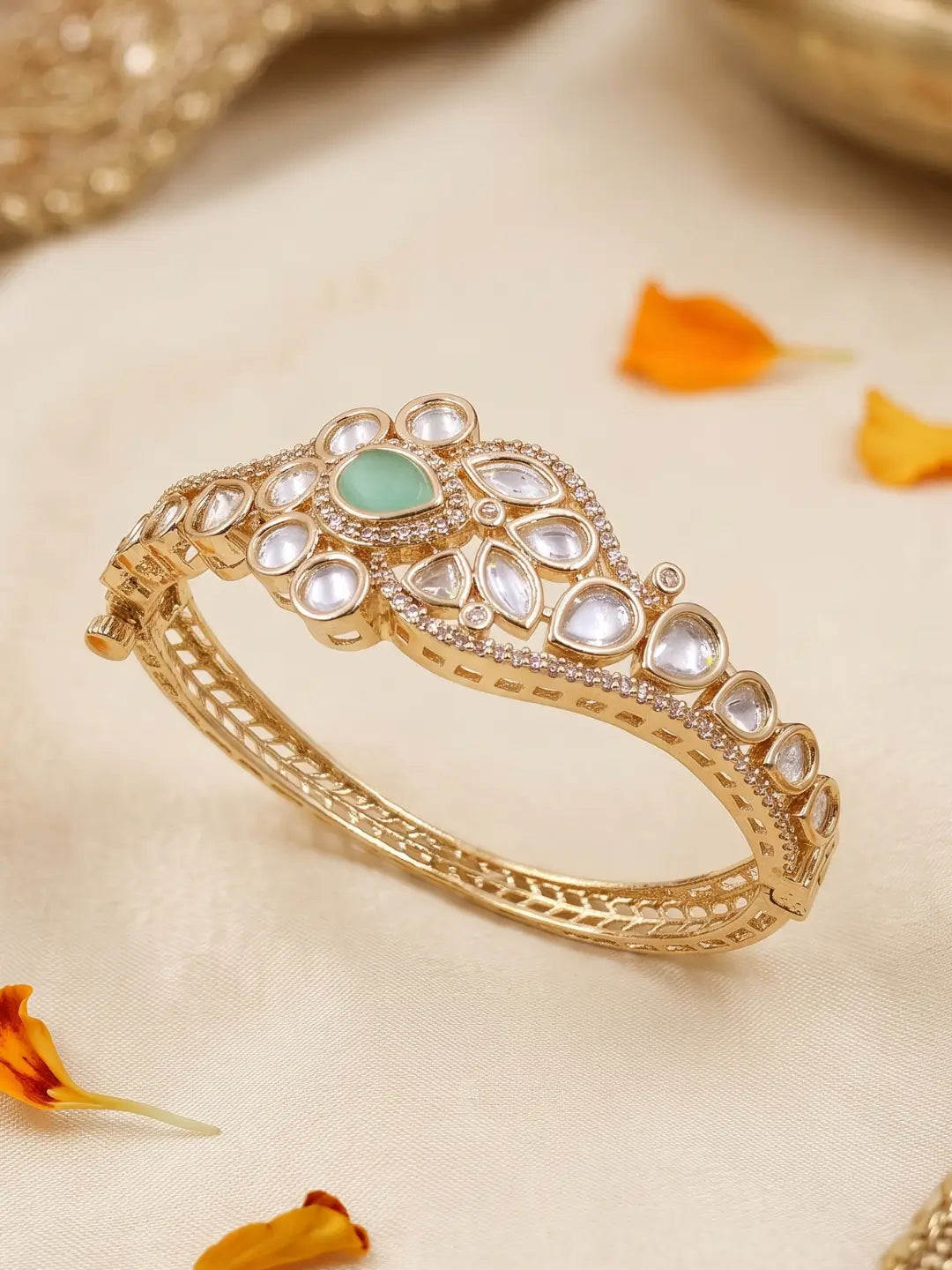 A gold-plated Kundan bangle bracelet with a central mint green stone and clear crystals, displayed on a cream-colored fabric with orange flower petals.