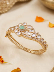 A gold-plated Kundan bangle bracelet with a central mint green stone and clear crystals, displayed on a cream-colored fabric with orange flower petals.