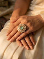 Close-up of a hand wearing an ornate gold-plated kundan statement ring with a mint green gemstone on a beige fabric background