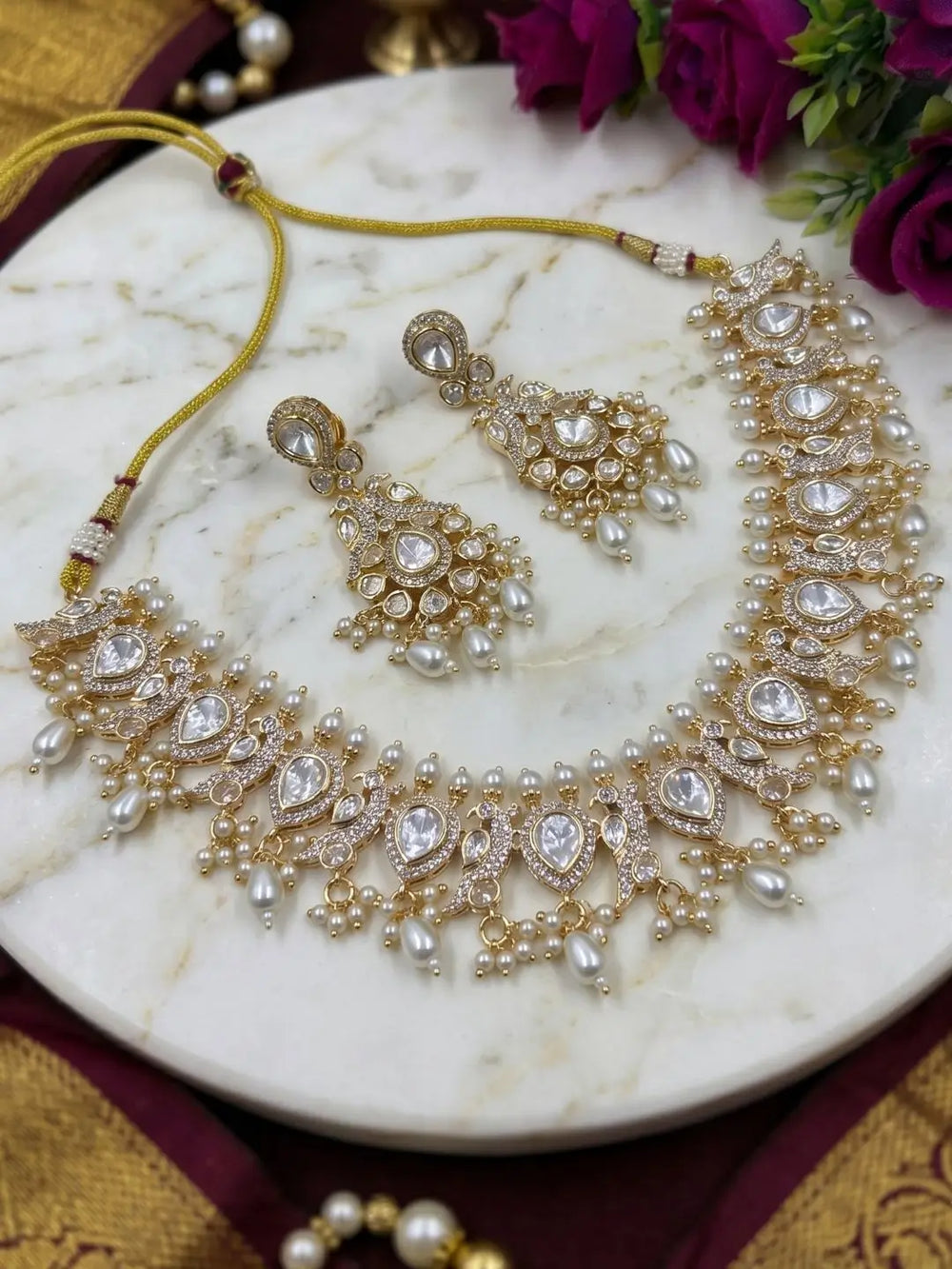 Gold and white pearl traditional Indian polki jewelry set, including a statement necklace, matching earrings, and a bracelet, displayed on a white marble plate with decorative flowers.