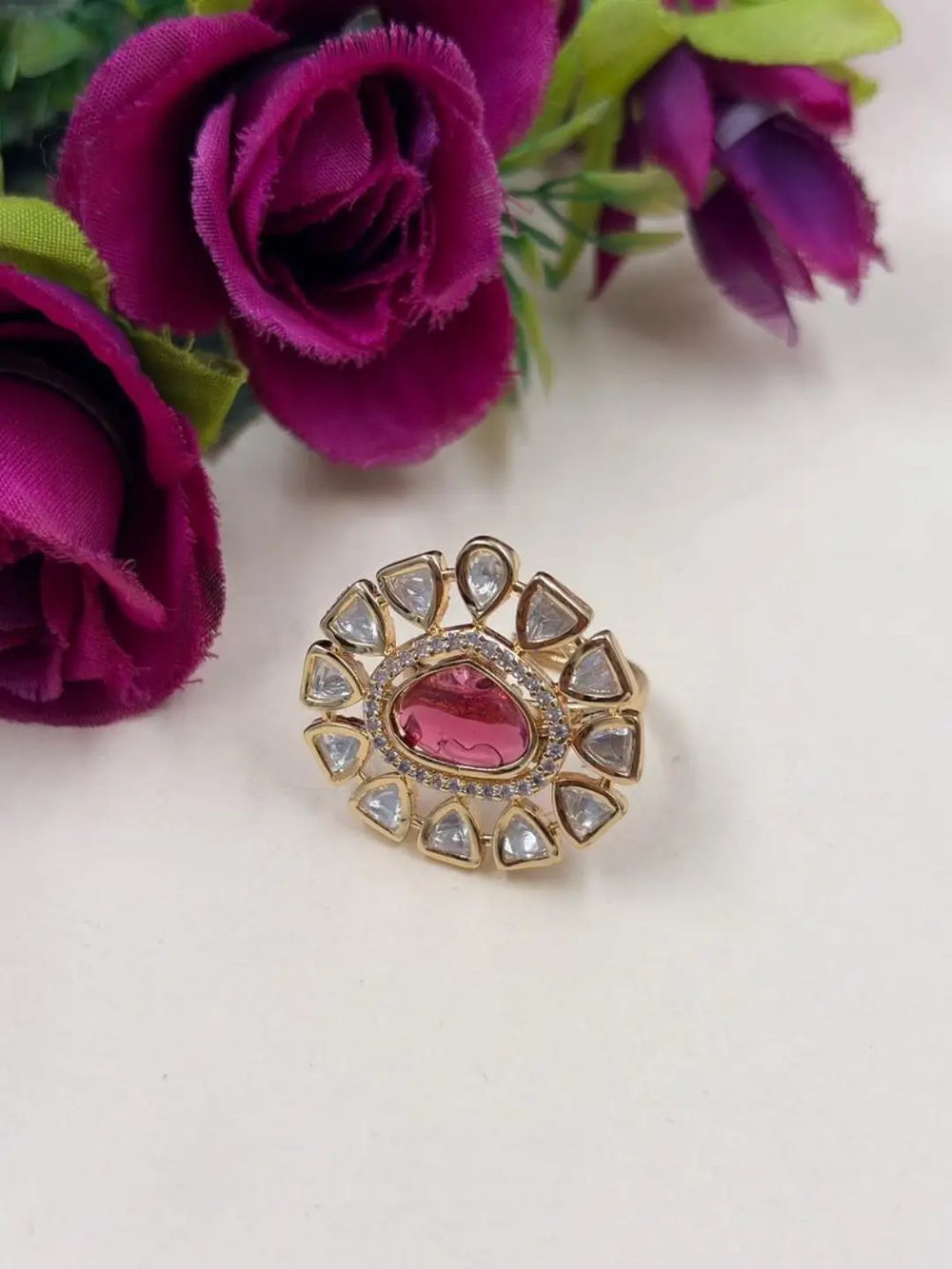 A gold-plated ring with a central pink stone and surrounding clear polki stones, styled on a white surface with purple flowers in the background.