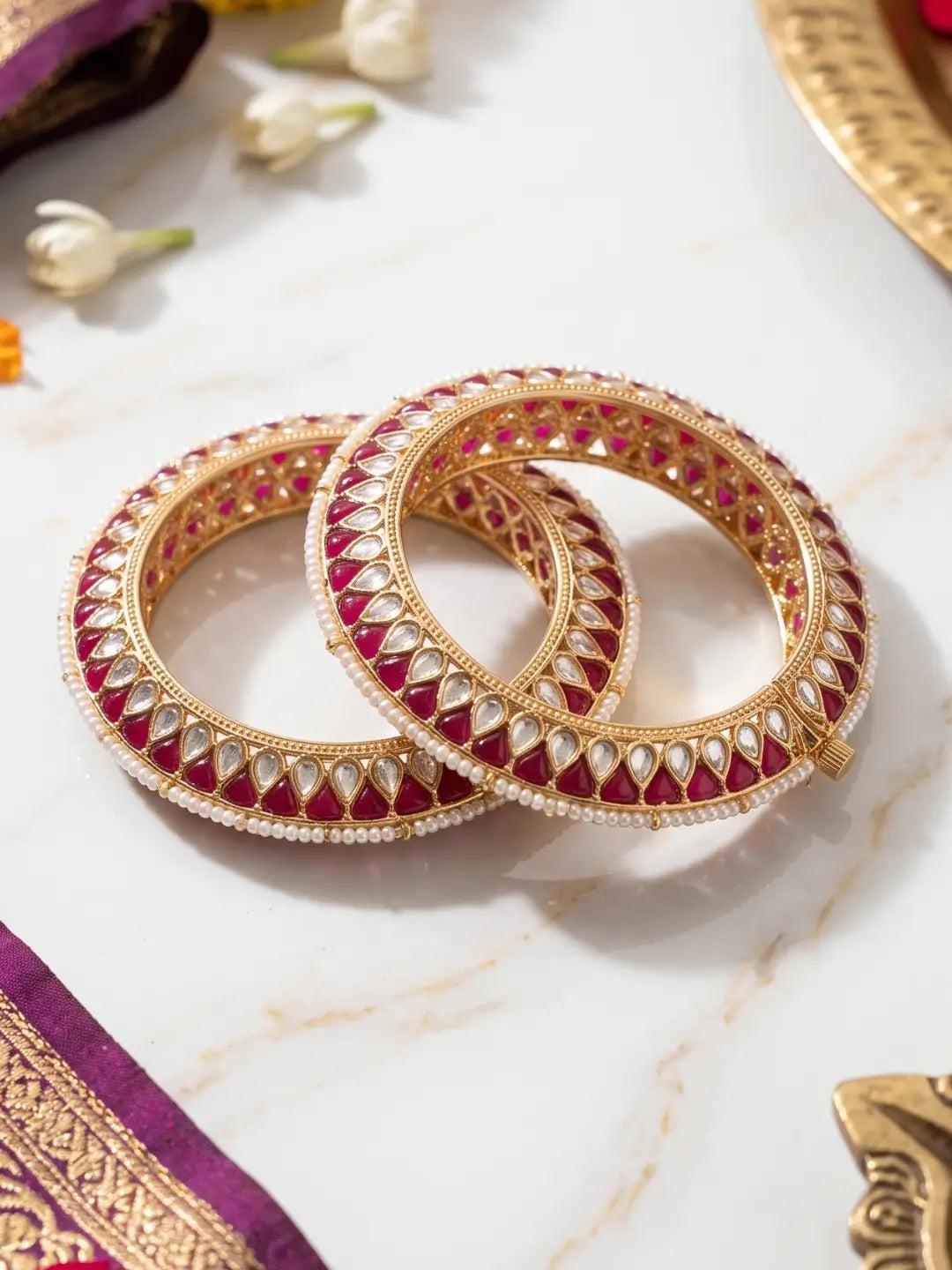 Two ornate gold bangles decorated with red enamel and white stones, arranged on a light marble surface with decorative floral and fabric props.