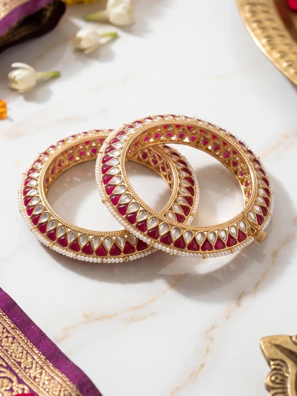 Two ornate gold bangles decorated with red enamel and white stones, arranged on a light marble surface with decorative floral and fabric props.