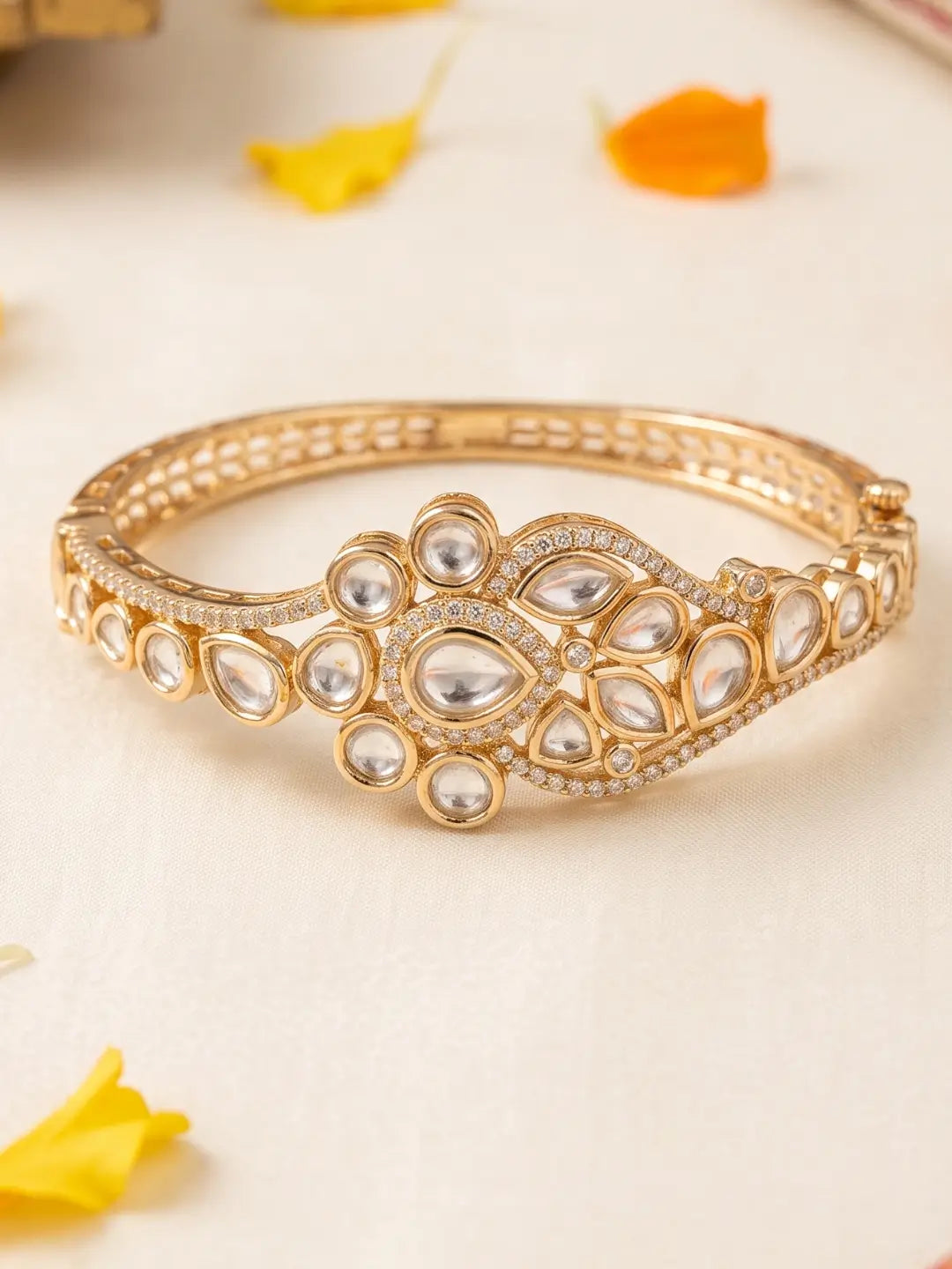 A gold-plated Polki bangle bracelet decorated with clear stone inlays, photographed on a light cream surface with a few scattered yellow and orange flower petals.