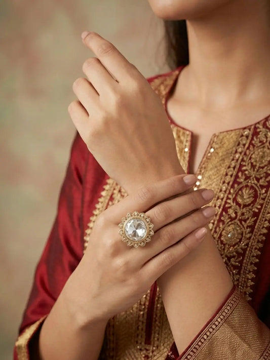 A close-up of a person wearing a large ornate, gold-plated kundan  ring with a clear central stone, styled with a deep red and gold embroidered traditional outfit.
