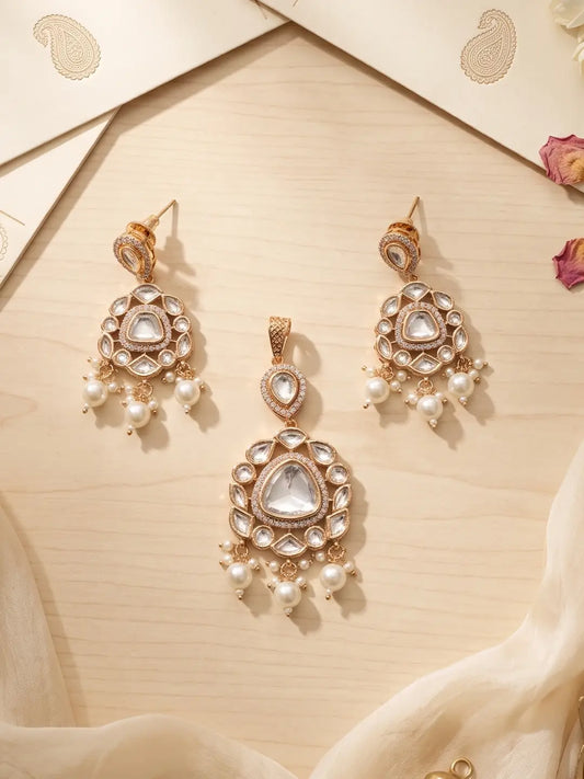 A matching gold-toned traditional Indian jewelry set featuring a pendant and two matching earrings, embellished with clear stones and white pearl beads, arranged on a light wood surface with decorative paper and fabric.