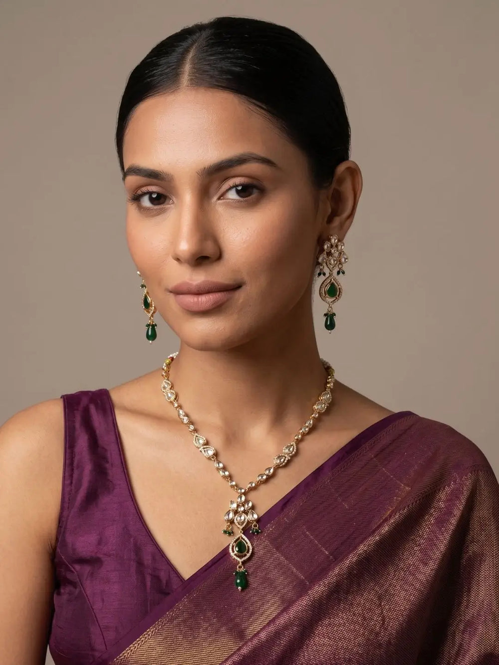 A woman wearing a gold-plated traditional kundan pendant necklace and matching drop earrings with green stone accents, styled with a deep purple saree against a neutral beige background.