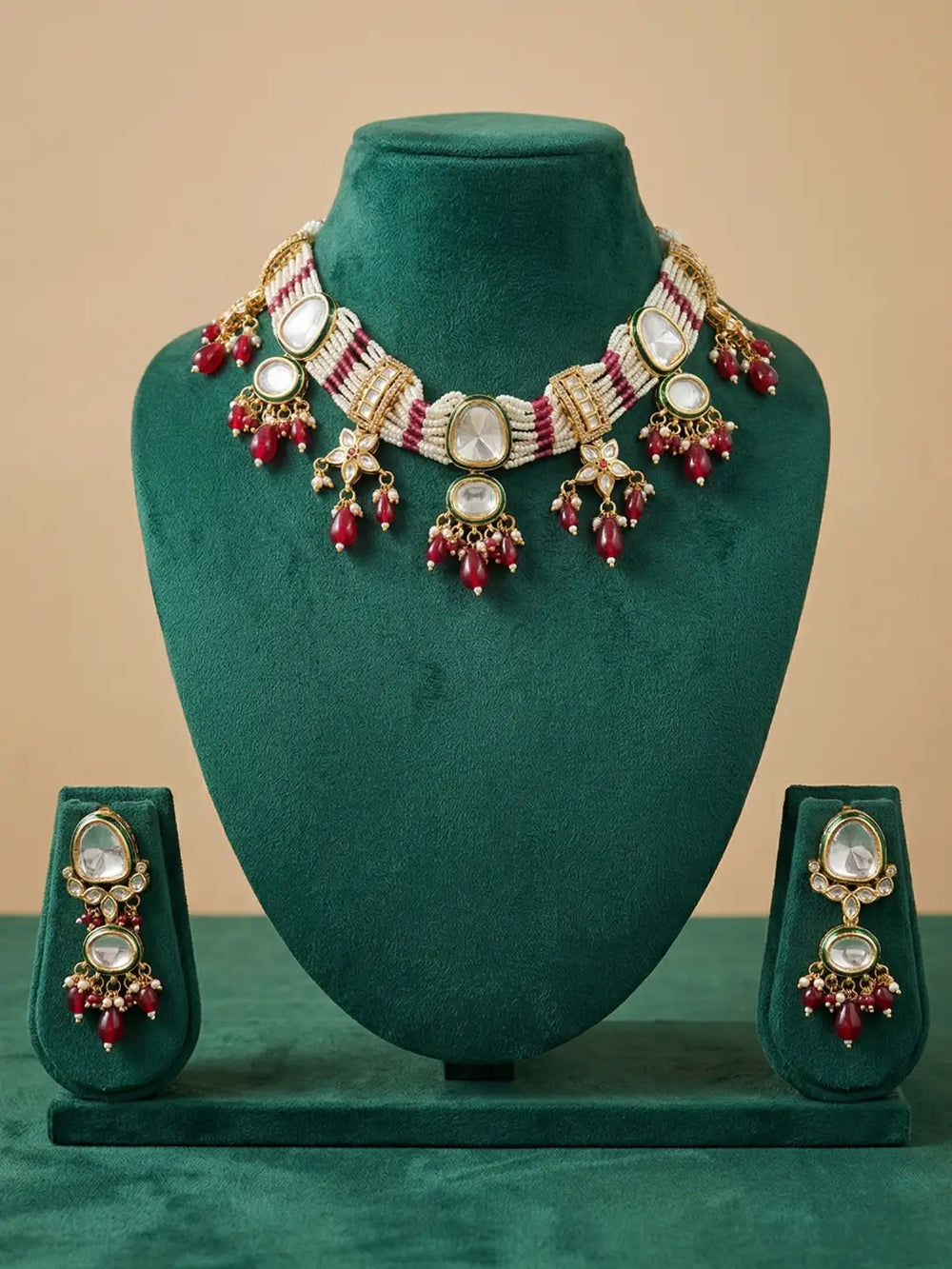 Gold-plated traditional Indian choker jewelry set featuring a statement choker necklace and matching earrings, embellished with white stones and red beads, displayed on a dark green velvet stand against a beige background.