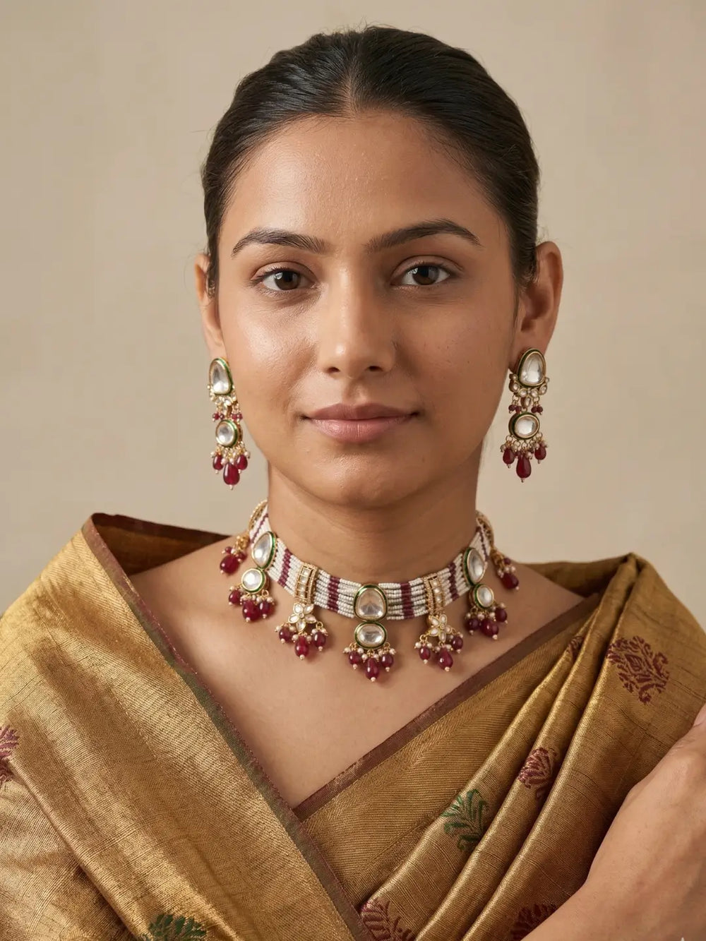 A woman wearing a traditional Indian gold saree with red and green floral patterns, styled with a gold-plated choker necklace and matching drop earrings with red stones and white pearls, set against a plain beige background.