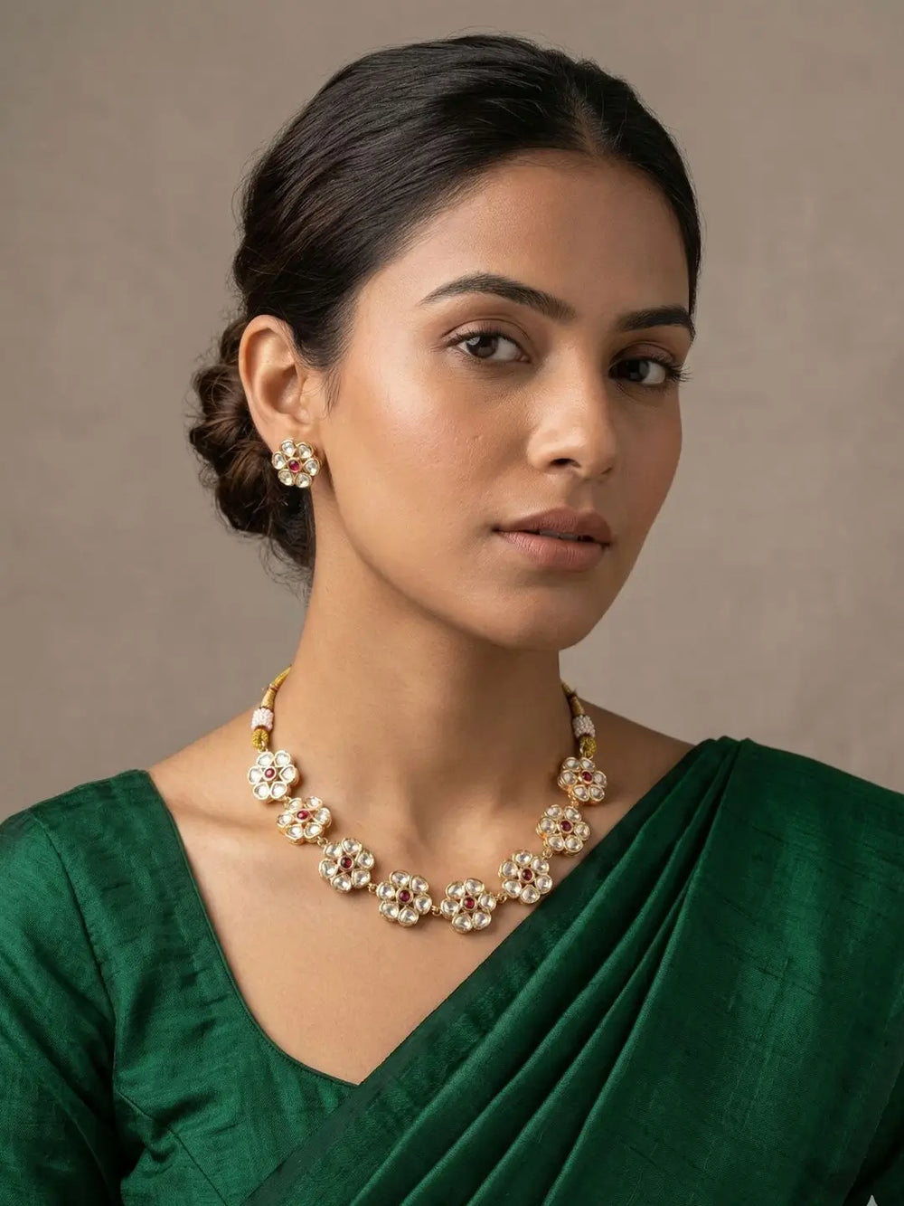 A woman wearing a gold-plated statement flower design polki necklace and matching stud earrings with clear stones, paired with a deep green sari, set against a neutral beige background.