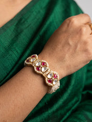 A close-up of a person's wrist wearing a gold-plated traditional Indian kundan polki and pearl bracelet adorned with red stones and clear stones, set against a dark green silk fabric.