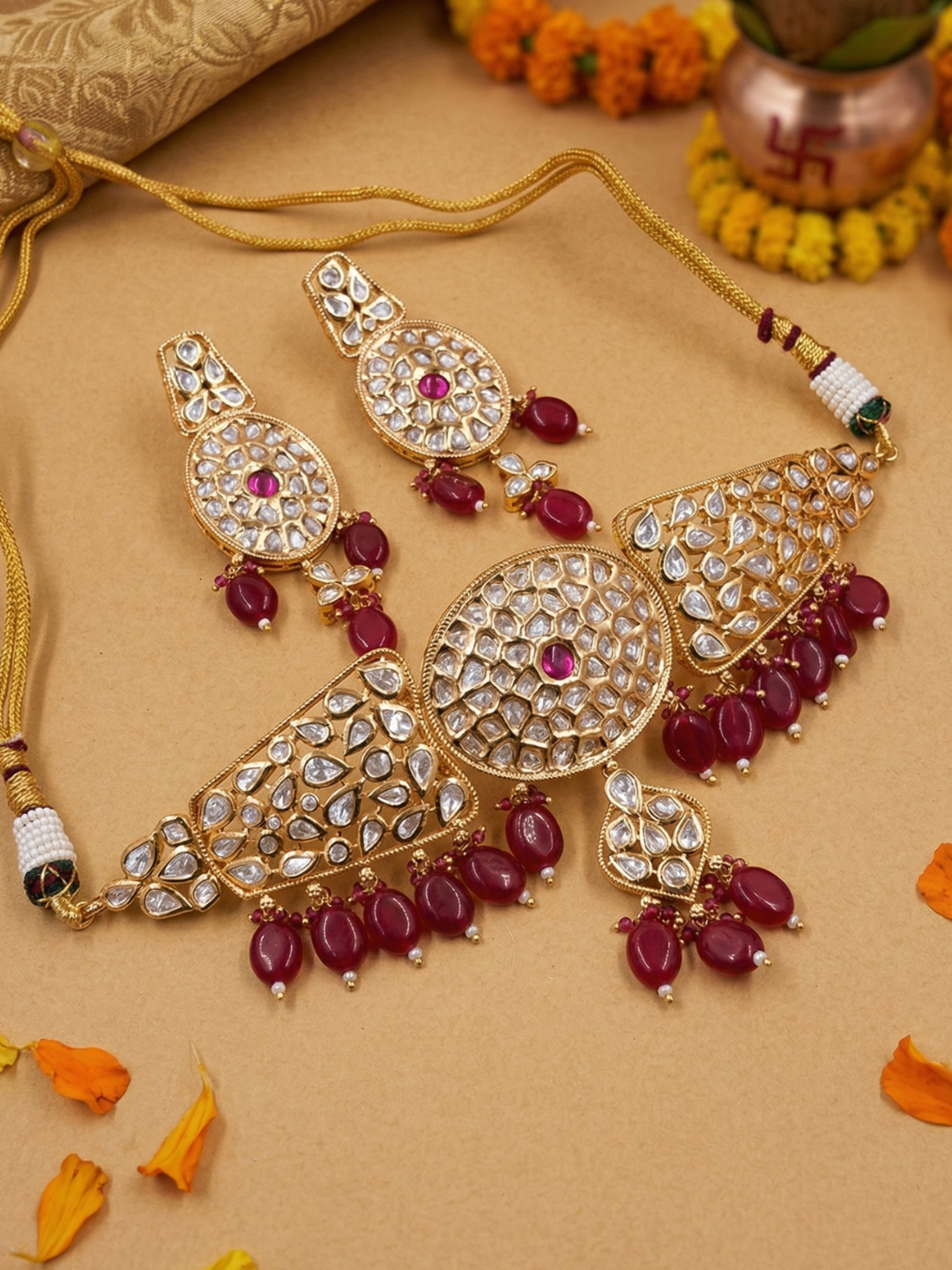 Gold-toned traditional Indian choker jewelry set featuring a necklace and matching earrings adorned with clear polki stones and deep red bead drops, arranged on a beige surface with decorative orange flower petals and a copper pot in the background.