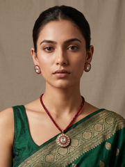 A woman wearing a green sari with gold zari embroidery, a red beaded necklace with a large gold-plated kundan pendant, and matching red stud earrings posed against a neutral beige background.