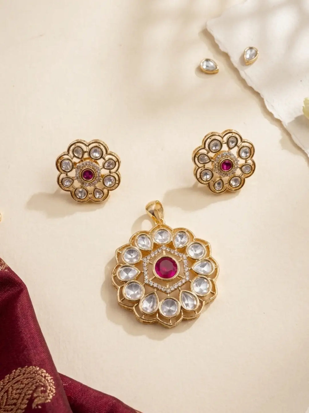 Gold-toned Indian kundan jewelry set featuring a large pendant, matching earrings, and a small stud, decorated with clear kundan stones and a central pinkish-red stone, arranged on a cream surface with decorative fabric.
