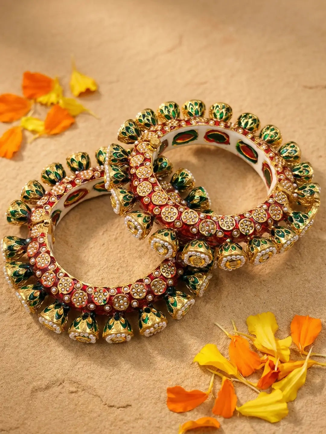 Two ornate Indian Kundan bangles with red, green, and gold hand-painted enamel and stone embellishments, displayed on a textured beige surface with orange and yellow flower petals.