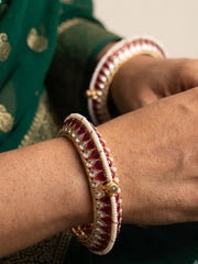 A close-up of a person's wrist wearing a traditional Indian bangle (kara) decorated with deep red enamel, white stones, and gold-plated borders, alongside a matching bangle on another wrist.