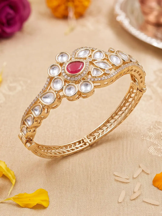 A gold-plated Kundan bangle bracelet decorated with clear crystals and a central red teardrop stone, displayed on a beige fabric background with decorative flower petals.