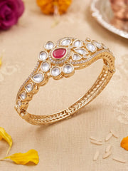 A gold-plated Kundan bangle bracelet decorated with clear crystals and a central red teardrop stone, displayed on a beige fabric background with decorative flower petals.