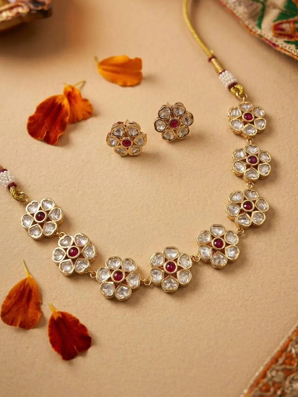 Gold-toned floral choker necklace and matching stud earrings with clear rhinestones and small red center stones, arranged on a beige surface with orange petal props.