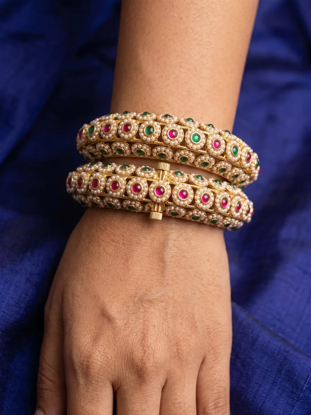 A close-up of a person's wrist and hand wearing stacked gold bangles adorned with pearl stones, red and green accents, set against a deep blue fabric background.
